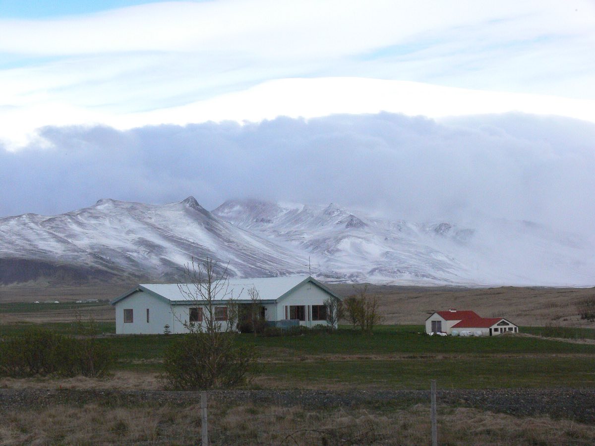 Bóndabær undir snævi þöktu fjalli, lágskýjað.